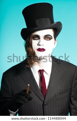 Gothic vampire looking business man wearing black striped suit and dark red tie. Another kind. Scary white face. Black vintage hat. Smoking cigar. Isolated on light blue background. Studio shot.