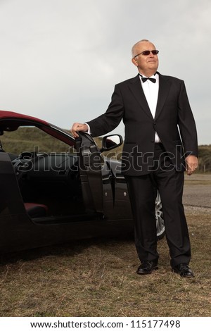 Senior good looking happy relaxed retired man with his sports car in dune landscape with cloudy sky. Wearing smoking suit and glasses. Classy different man.