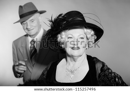 Good looking senior couple glamour vintage style. Wearing a hat. Black and white studio shot. Short blonde curly hair. Chic look. Dressed in black. Wearing a grey suit with tie.