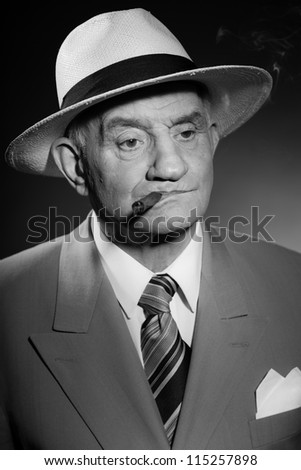 Senior glamour vintage man wearing suit and tie and hat. Black and white studio shot. Gangster look. Smoking cigar and drinking glass of whisky. Isolated.