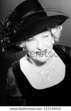 Good looking senior woman glamour vintage style. Wearing a black hat. Black and white studio shot. Short blonde curly hair. Chic look. Dressed in black.