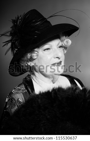 Good looking senior woman glamour vintage style. Wearing a black hat. Holding a black fan. Black and white studio shot. Short blonde curly hair. Chic look. Dressed in black.