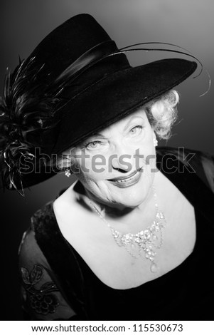 Good looking senior woman glamour vintage style. Wearing a black hat. Black and white studio shot. Short blonde curly hair. Chic look. Dressed in black.