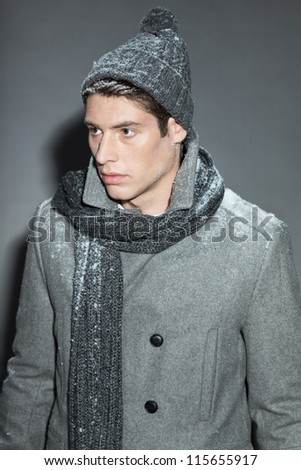 Men winter fashion. Handsome man with brown hair wearing grey scarf, grey woolen hat and grey coat. Covered with snow. Cold. Casual look. Studio shot isolated on grey background.