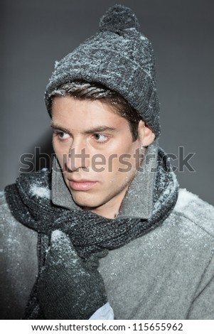 Men winter fashion. Handsome man with brown hair wearing grey scarf, grey woolen hat, grey woolen gloves and grey coat. Covered with snow. Cold. Casual look. Studio shot isolated on grey background.