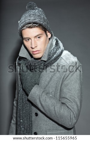 Men winter fashion. Handsome man with brown hair wearing grey scarf, grey woolen hat, grey woolen gloves and grey coat. Covered with snow. Cold. Casual look. Studio shot isolated on grey background.