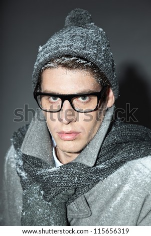 Men winter fashion. Handsome man with brown hair wearing grey scarf, grey woolen hat, grey woolen gloves grey coat and black glasses. Covered with snow. Cold. Casual look. Studio shot isolated on grey