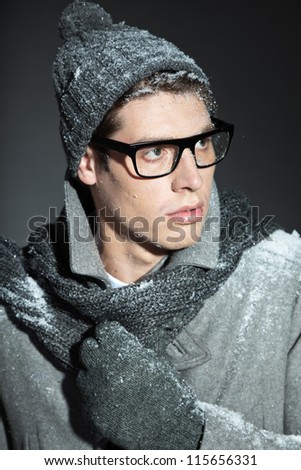 Men winter fashion. Handsome man with brown hair wearing grey scarf, grey woolen hat, grey woolen gloves grey coat and black glasses. Covered with snow. Cold. Casual look. Studio shot isolated on grey