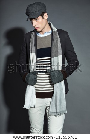 Men winter fashion. Handsome man wearing grey scarf, striped sweater, grey jacket, white pants, grey woolen cap and gloves. Casual look. Studio shot isolated on grey background.