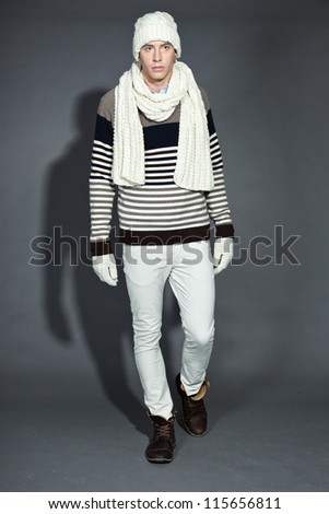 Men winter fashion. Handsome man wearing white scarf, striped sweater, white pants, white woolen hat and gloves. Casual look. Studio shot isolated on grey background.