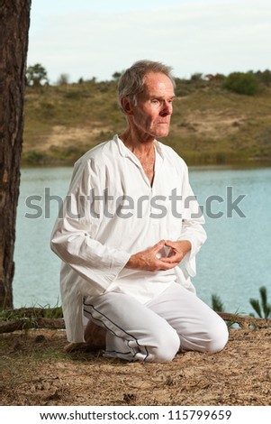 Senior spiritual man dressed in white. Short grey hair. Meditating in nature. Outdoors. Forest. Dunes. Cloudy sky. Healthy living.