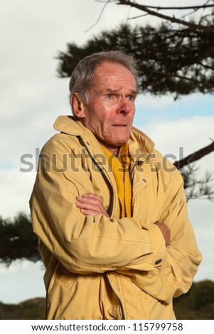 Senior man with yellow coat. Having cold. Sad looking. Warming up. Short grey hair. Autumn. Outdoors. Dune landscape. Cloudy sky. Healthy living.