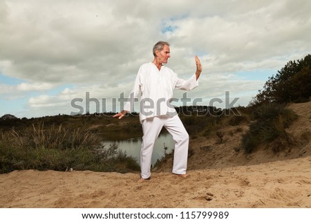 Senior spiritual man dressed in white. Short grey hair. Working out in nature. Outdoors. Dune landscape. Cloudy sky. Healthy living.