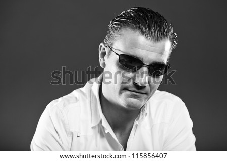 Macho handsome man with short brown hair wearing white shirt and sunglasses. Mafia type. Good looking. Fashion studio portrait. Isolated on grey background. Glamour black and white portrait.