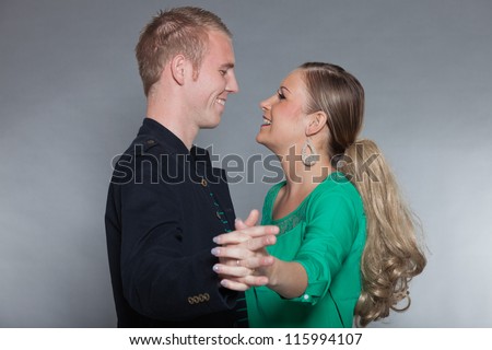 Happy young couple dancing together. Cool looking. Chic dressed. Dark jacket. Green shirt. Jeans. Man short blonde hair. Woman long brown hair. Studio shot isolated on grey background.