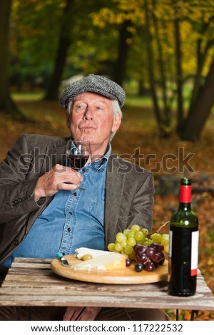 Senior french man enjoying red wine and cheese outdoors in autumn forest.