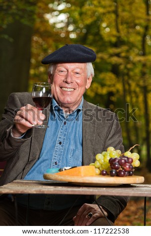 Senior french man enjoying red wine and cheese outdoors in autumn forest.
