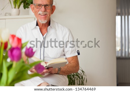 Senior man with glasses reading book in living room.