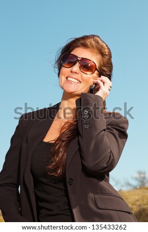 Pretty brunette woman with sunglasses using mobile phone. Meadow with blue sky.