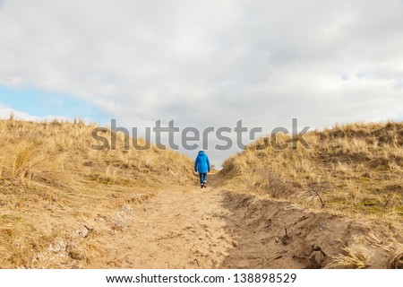Tourist walking on path in grassy dune landscape with cloudy sky.