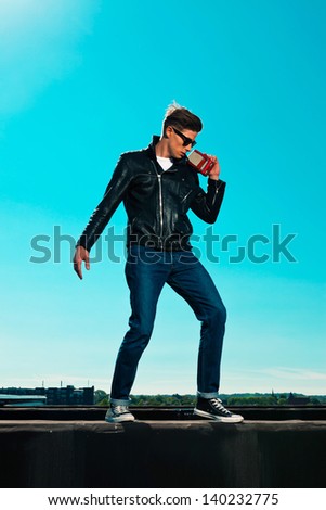 Rockabilly man retro 50s style with black jacket listens to portable radio. On rooftop. Blue sky.
