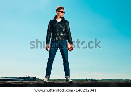 Rock and roll man 50s style with black jacket. Holding portable radio. On rooftop. Blue sky.