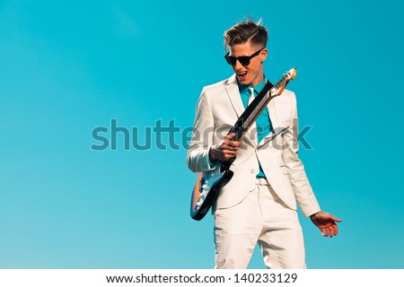 Retro fifties male electric guitar player wearing white suit and sunglasses. Blue sky.