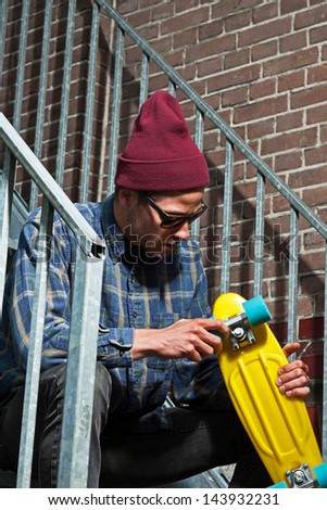 Urban skateboarder with woolen hat and sunglasses holding his board sitting on iron stairs.
