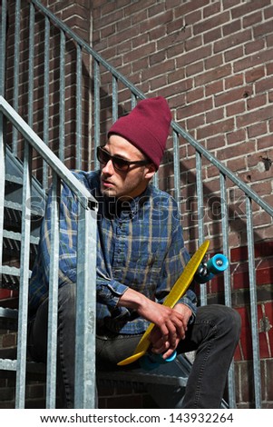 Urban skateboarder with woolen hat and sunglasses holding his board sitting on iron stairs.