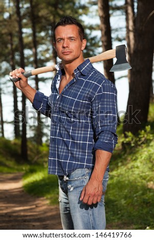 Handsome strong lumberjack man holding axe. Wearing blue shirt. Standing in forest.