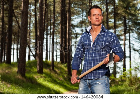 Handsome strong lumberjack man holding axe. Wearing blue shirt. Standing in forest.