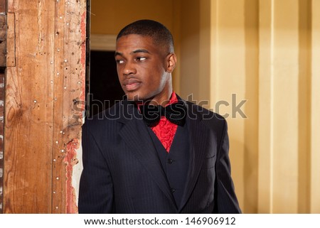 Retro fashion afro american groom wearing black suit and tie and red tie. In doorpost of an old house.