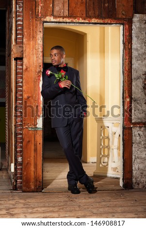 Vintage fashion afro american groom holding red rose. Standing in doorpost of an old house.