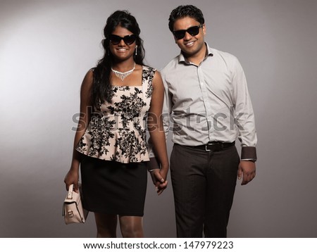 Fashionable indian couple walking towards camera. Wearing retro black sunglasses. Studio shot against grey.