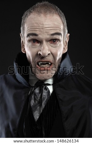 Dracula with black cape showing his scary teeth. Vamp fangs. Studio shot against black.
