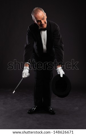 Magician with black suit and hat holding a magic stick. Studio shot against black.