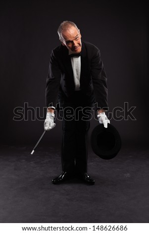 Magician with black suit and hat holding a magic stick. Studio shot against black.