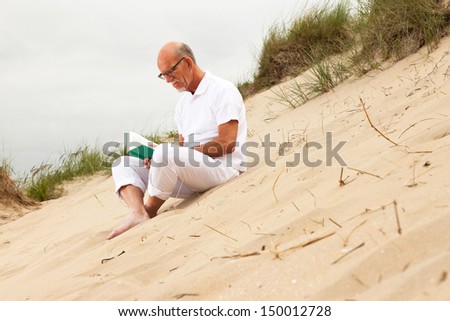 Retired man with beard and glasses reading a book in grass dune landscape. Dressed in white.