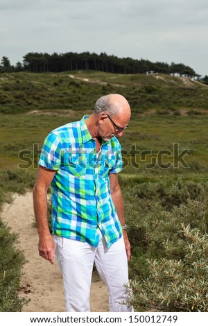 Retired senior man walking outdoors in grass dune landscape. Wearing white pants and green blocked shirt.