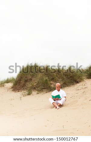 Retired man with beard and glasses reading a book in grass dune landscape. Dressed in white.