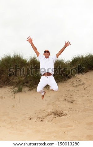 Jumping happy retired man with beard and sunglasses in grass dune landscape with cloudy sky.
