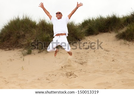 Jumping happy retired man with beard and sunglasses in grass dune landscape with cloudy sky.