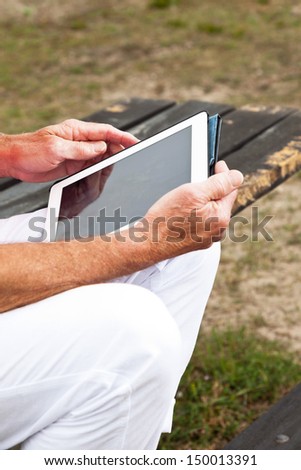 Close-up of hands of retired senior man resting and using his tablet at table in grass dune landscape.