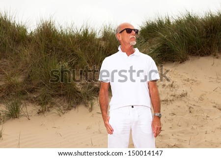 Retired man with beard and sunglasses in grass dune landscape with cloudy sky. Dressed in white.