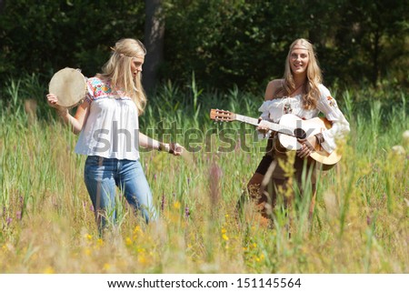 Two retro blonde 70s hippie girls making music with acoustic guitar and tambourine outdoor in nature.