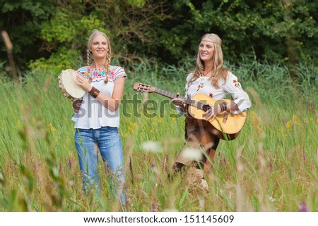 Two retro blonde 70s hippie girls making music with acoustic guitar and tambourine outdoor in nature.