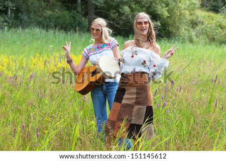 Two retro blonde 70s hippie girls with sunglasses making music with acoustic guitar and tambourine outdoor in nature.