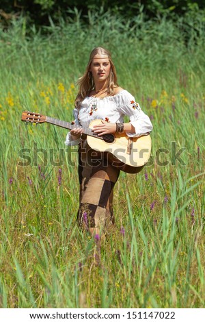 Retro blonde 70s hippie girl with acoustic guitar outdoor in nature. Standing in meadow.
