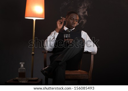 Retro african american gangster man wearing striped suit and tie. Sitting in a chair in living room. Smoking cigar. Holding glass of whisky.