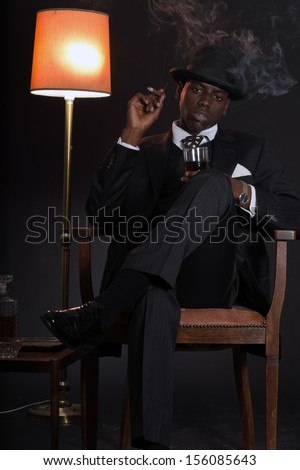 Retro african american gangster man wearing striped suit and tie and black hat. Sitting in a chair in living room. Smoking cigar. Holding glass of whisky.